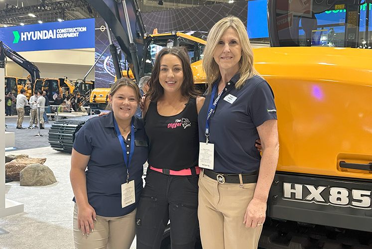 Amy Underwood stands between two women in front of a yellow HX85A Hyundai construction vehicle at CONEXPO-CON/AGG