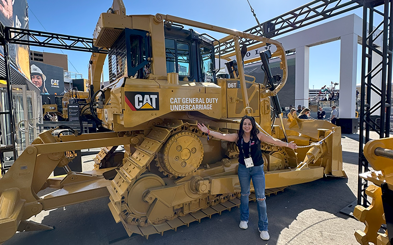 Amy standing with her arms out in front of a CAT bulldozer at CONEXPO-CON/AGG
