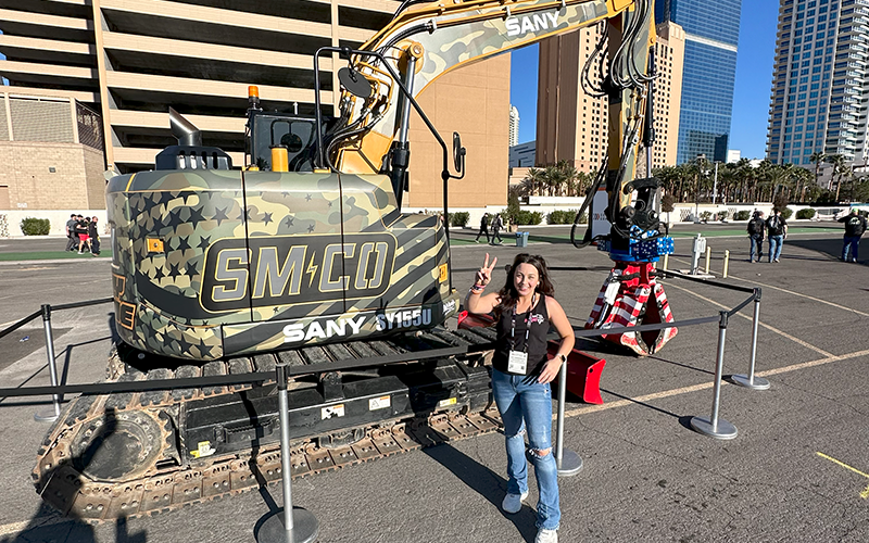 Amy Underwood posing with a peace sign in front of SM-CO SANY Excavator at CONEXPO-CON/AGG