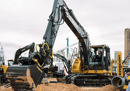 John Deere excavator with Engcon bucket full of sand at a busy construction site, with other vehicles, cranes, and buildings in the background