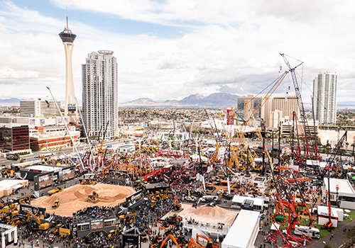 Outdoor construction expo with cranes, excavators, and crowds, set in a city with tall buildings, a Stratosphere-like tower, distant mountains, and a partly cloudy sky