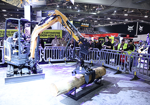 Mini excavator with yellow arm lifting a large log at CONEXPO-CON/AGG, surrounded by metal barriers and onlookers