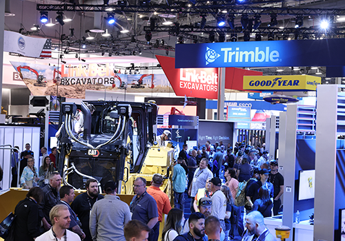 Busy CONEXPO-CON/AGG with booths and banners for Trimble, Link-Belt, and Goodyear. A crowd gathers around a Caterpillar machine.