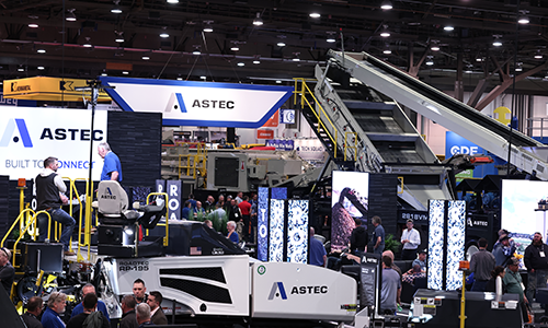ASTEC booth at CONEXPO-CON/AGG with heavy machinery, a large conveyor system, digital screens, and the slogan ‘BUILT TO CONNECT.’ Attendees explore equipment and engage with exhibits