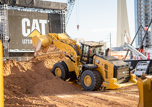 Yellow CAT bulldozer moving dirt at a construction site, with buildings, cranes, and a large 'CAT LET'S DO' sign in the background