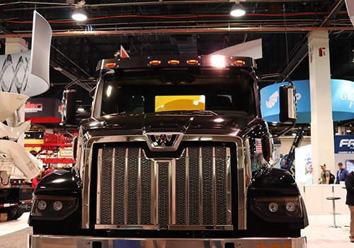 Front view of a large black truck with a chrome grille and a 'W' logo on the hood, showcased indoors at a trade show with banners and other vehicles in the background under bright lighting