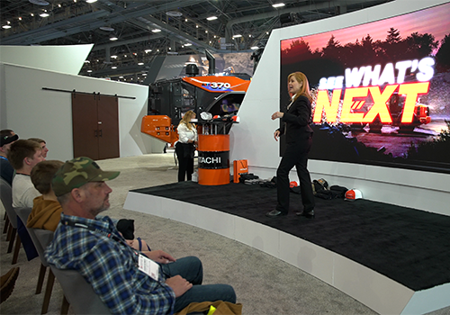 Presenter on stage at CONEXPO-CON/AGG with a large screen reading 'SEE WHAT'S NEXT', seated audience, and an orange Hitachi machine in the background.