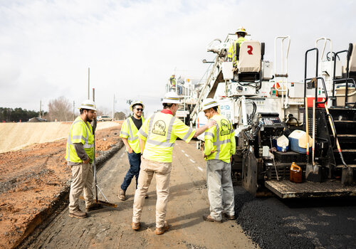A group of construction workers in safety vests and hard hats stand on a road under construction, gathered around paving machinery. The background shows more equipment and an unfinished road section.