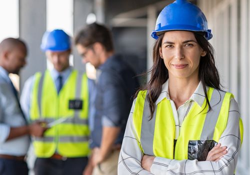 Women Construction worker with arms crossed and wearing blue hard hat, yellow vest and workers in the back