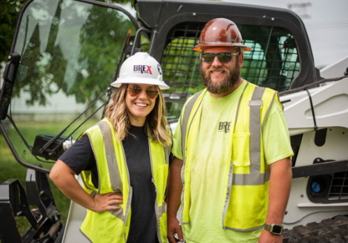 Guy and women in front of skid-steer machine in yellow vest and hard hat