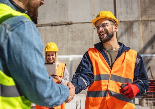 Three construction workers wearing safety vests and hard hats are at a construction site. Two of the workers are shaking hands, while the third worker is holding a clipboard in the background. The faces of all three workers are blurred out.