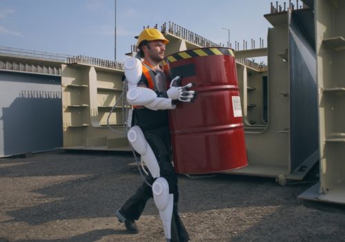 A person wearing a yellow hard hat and an exoskeleton suit is carrying a large red barrel with black and yellow caution tape on the top. The background shows industrial structures and equipment.