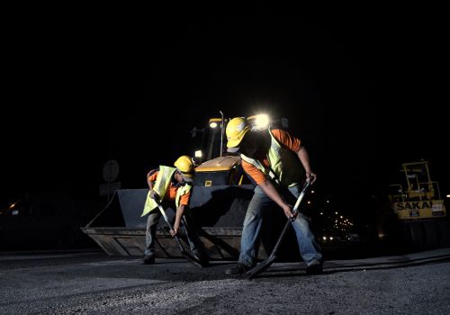 Two construction workers wearing yellow hard hats and reflective vests are working on a road at night. They are using shovels to spread asphalt, with a large piece of machinery behind them providing light. The background is dark, with some lights visible in the distance