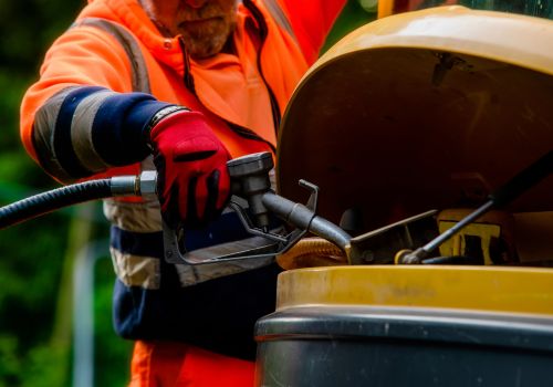 A person wearing an orange high-visibility jacket and red gloves is refueling a yellow construction vehicle. The fuel nozzle is inserted into the vehicle's fuel tank, and the person's hand is gripping the nozzle handle. The background shows greenery, indicating an outdoor setting.