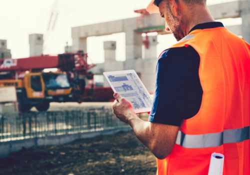 A construction worker in an orange safety vest and hard hat uses a tablet at an active construction site, with concrete structures, scaffolding, a crane, and a truck in the background.