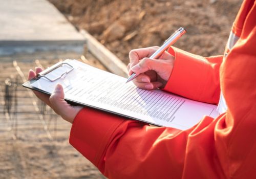 A person wearing an orange jacket is holding a clipboard and writing on a form with a pen. The background shows an outdoor construction site with dirt and concrete structures.