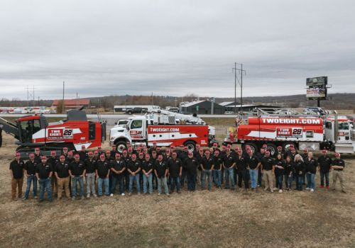 A large group of people wearing black shirts and jeans stand in front of several red and white trucks branded with 'TWOEIGHT.COM' and the number '29'. The background includes a cloudy sky, buildings, and a billboard.