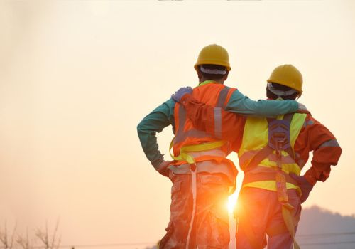 Two construction workers wearing hard hats and reflective safety vests stand side by side with their arms around each other, looking at the sunset. The sun is low in the sky, casting a warm glow on their backs.