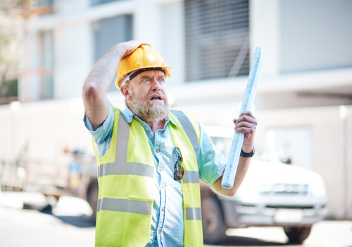 A mature building professional looks up, seeming worried and on the verge of panic at something he can see on his building site.