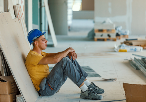 A young Caucasian construction worker is sitting down and resting with his eyes closed, due to feeling exhausted and dizzy.