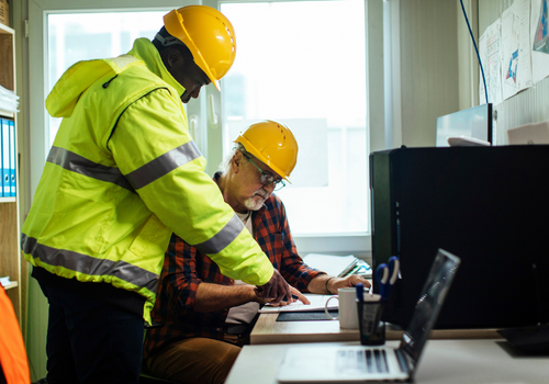 Two male engineers with the hardhats looking at the blueprints in their office at the construction site