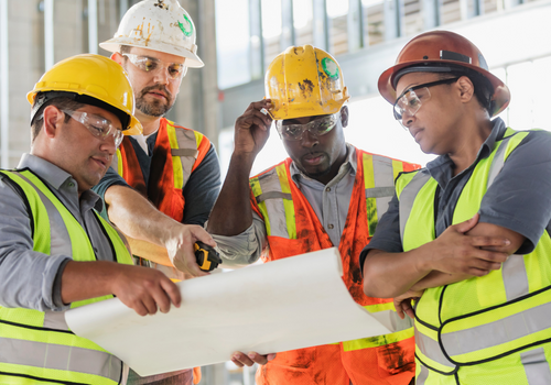 A multi-ethnic group of four construction workers wearing hardhats and safety vests, having a meeting inside the structure being built. A young Hispanic man is holding a set of floor plans which they are carefully studying. The worker on the right is a mature African-American woman in her 40s.