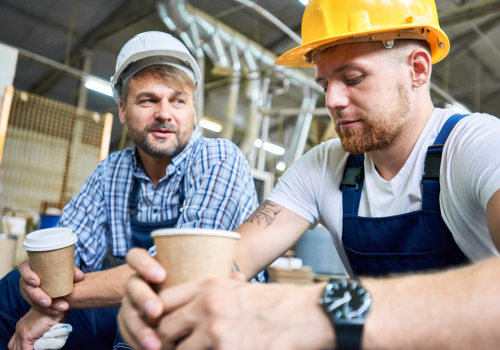 two workers wearing hardhats taking break from work drinking coffee and resting sitting on construction site